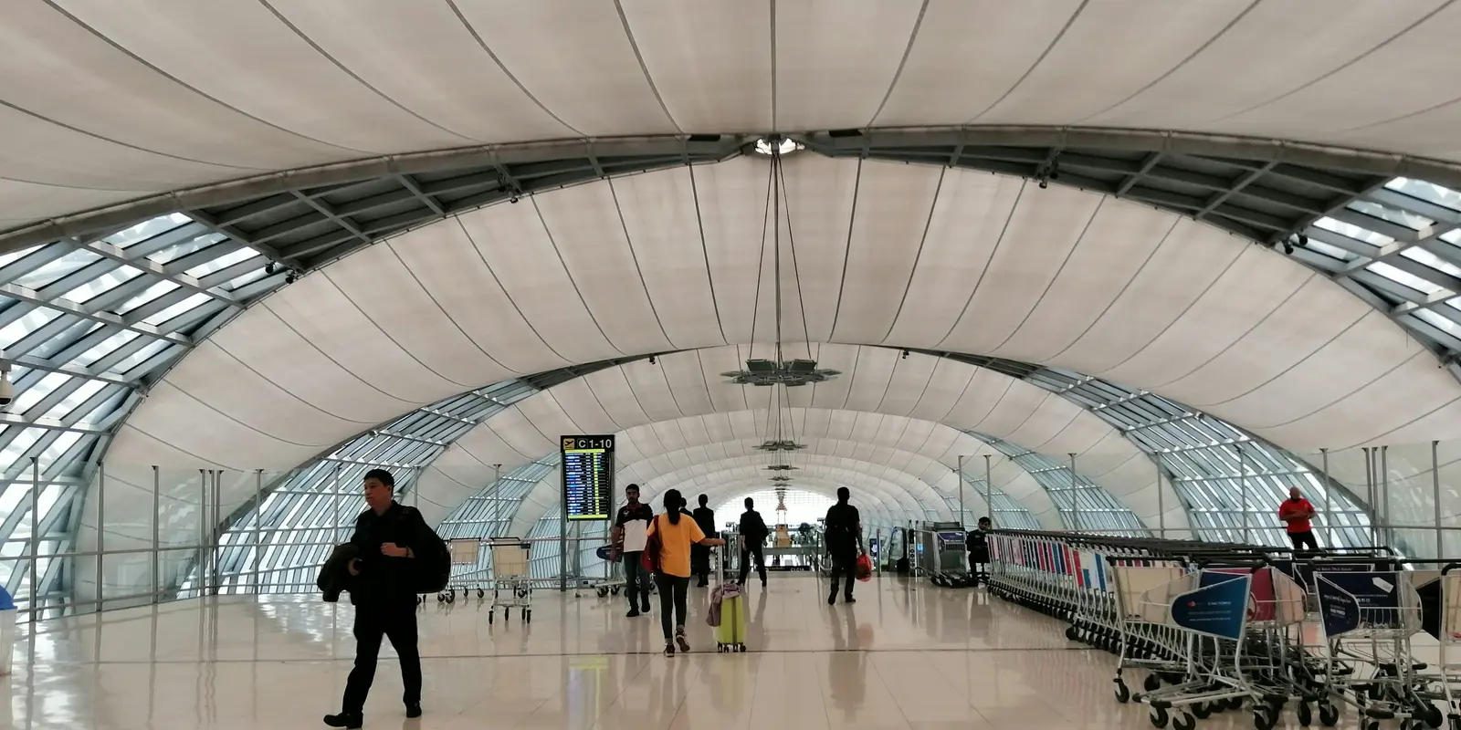 Travelers walking through a spacious airport terminal hall with a curved ceiling and a flight information board in the background